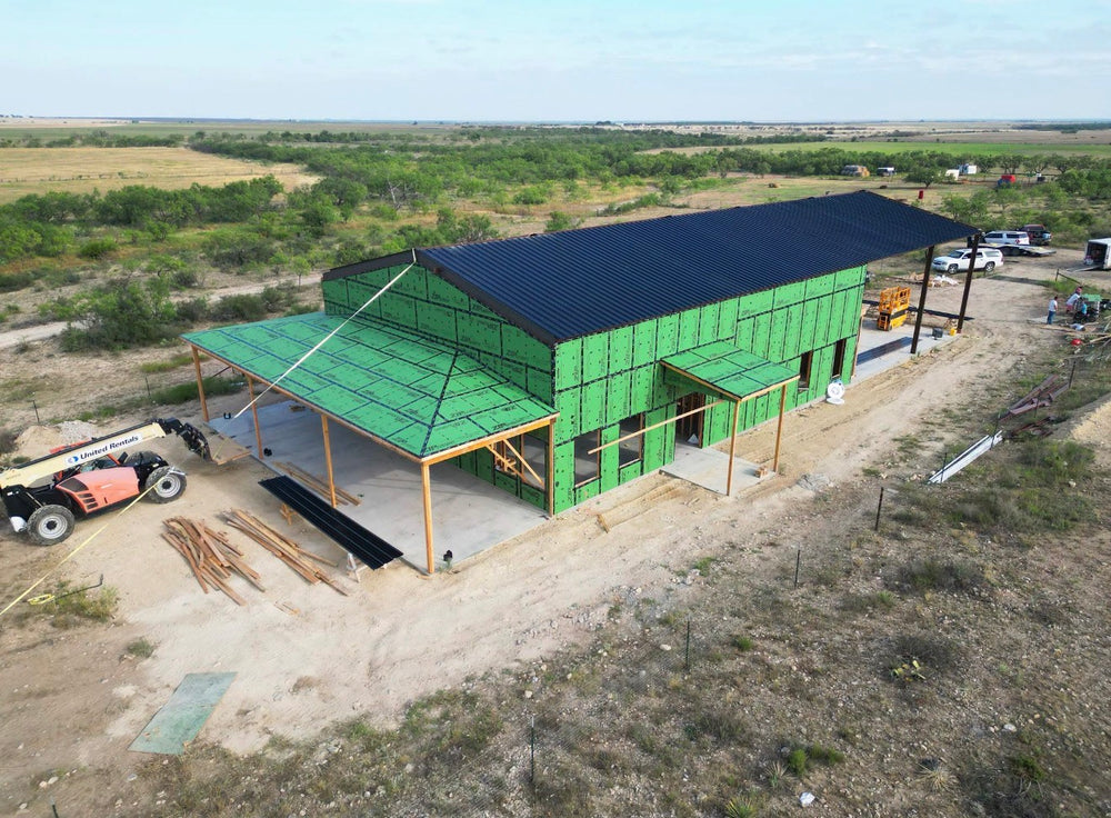 Aerial view of a barndominium under construction with green ZIP System sheathing, a black standing seam metal roof, and timber porch posts