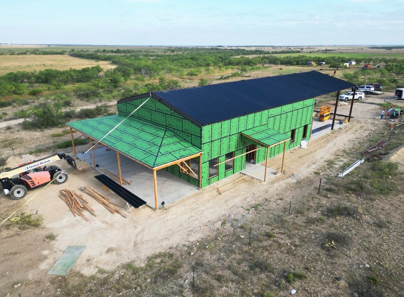 Aerial view of a barndominium under construction with a black metal roof and wraparound porch framing on open Texas ranch land