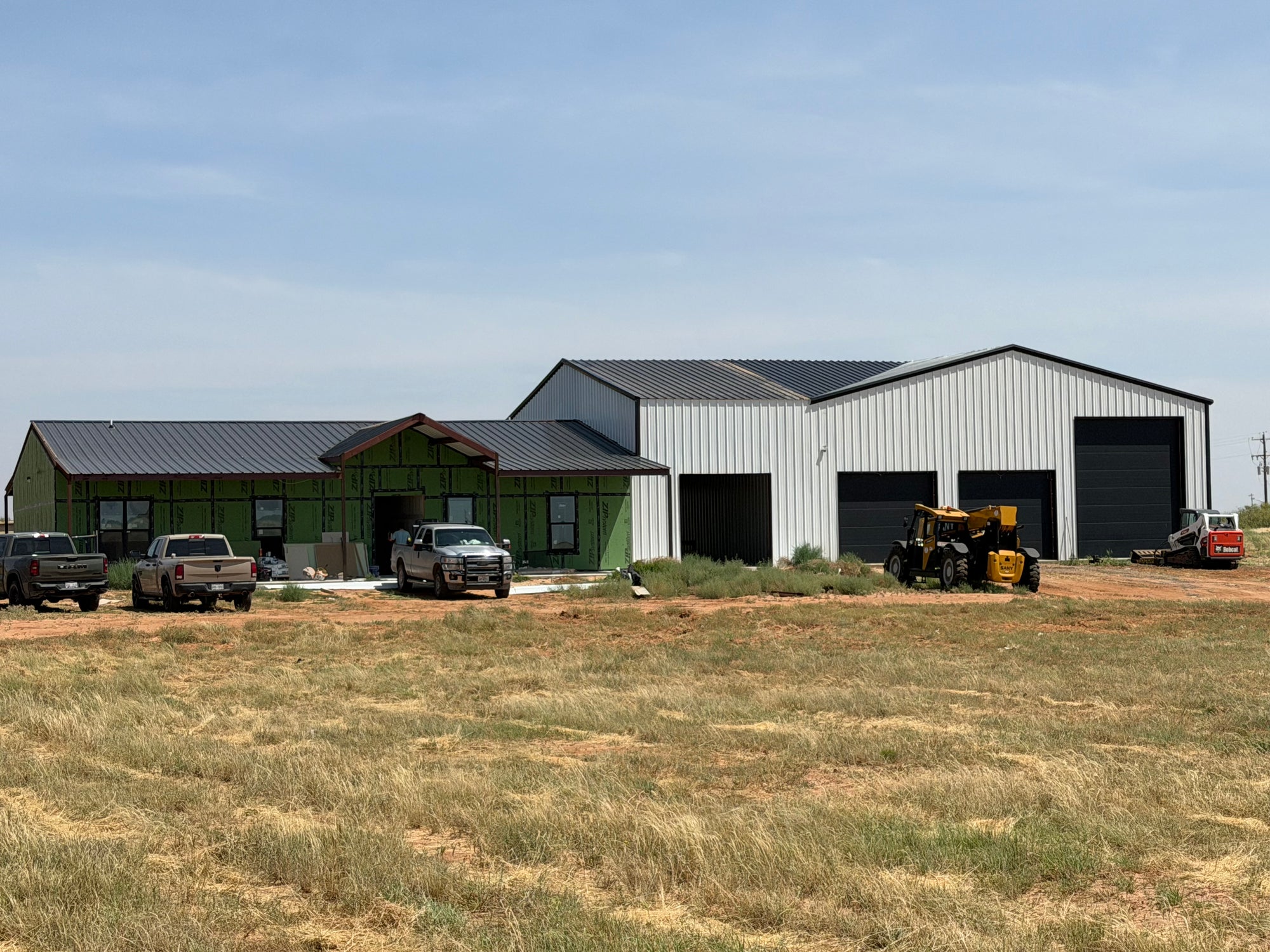 Barndominium residence and attached metal shop nearing completion, with contractor trucks and a telehandler parked on the job site