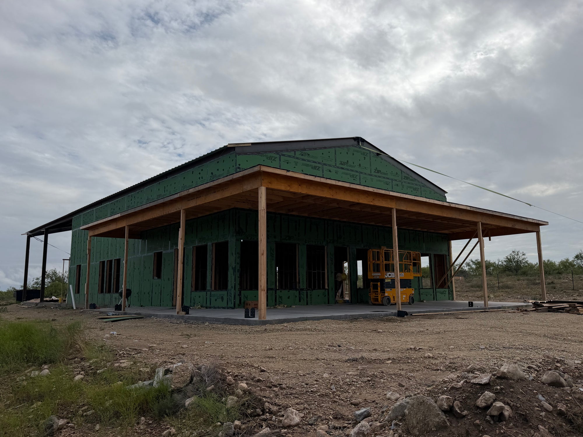 Barndominium under construction with green ZIP System sheathing, a deep wraparound covered porch, and a scissor lift on the slab