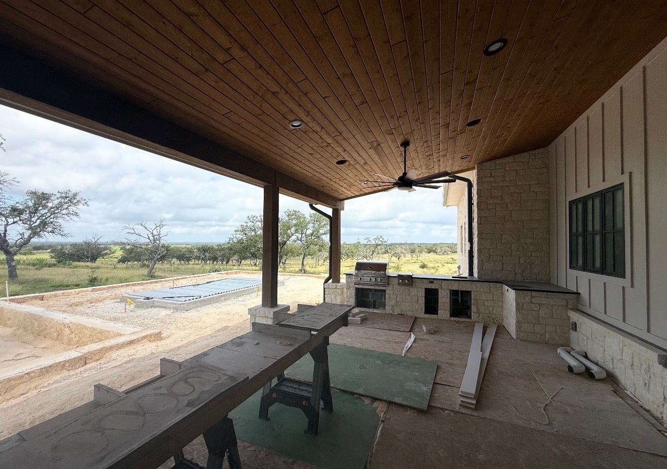 Covered outdoor living area with a stained wood plank ceiling, ceiling fan, outdoor kitchen under construction, and a pool beyond