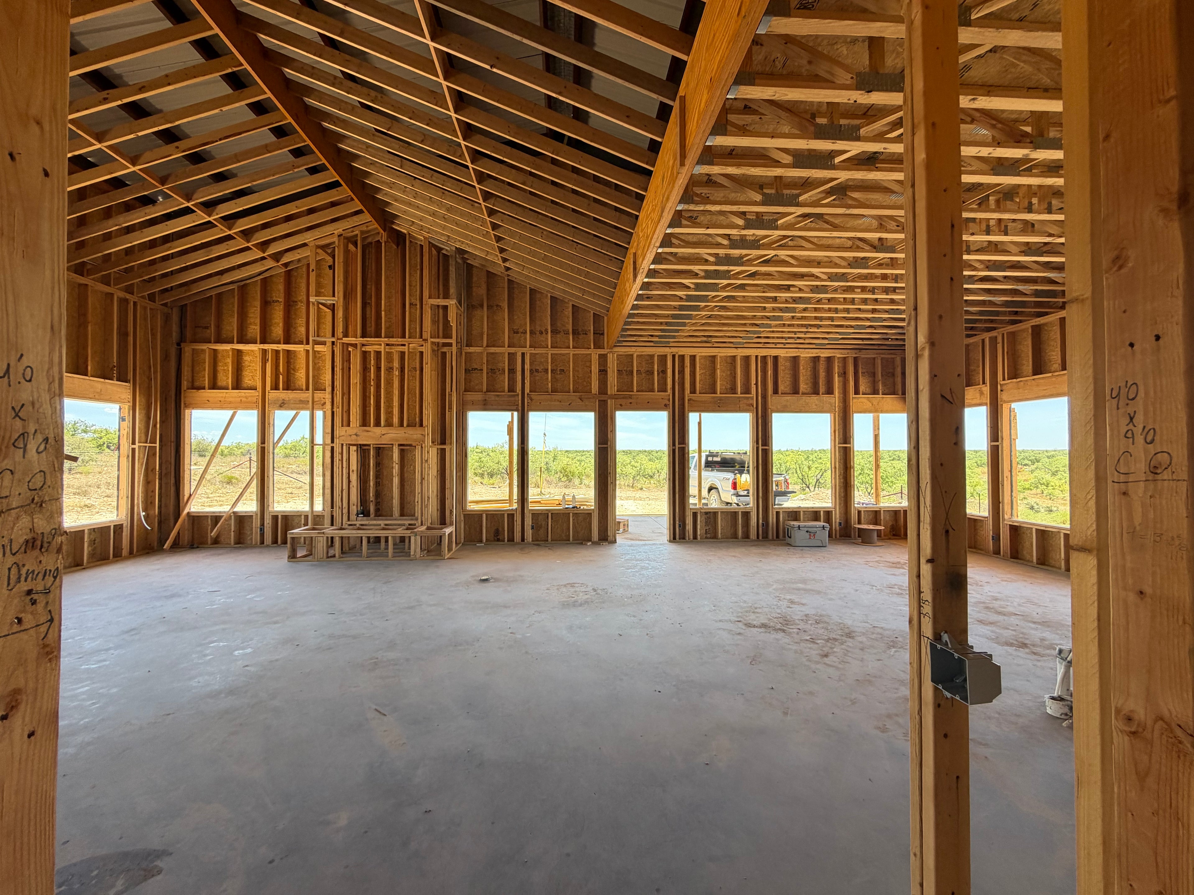 Interior framing of a custom home great room with a cathedral ceiling, exposed rafters, and a wall of window openings overlooking Texas brush