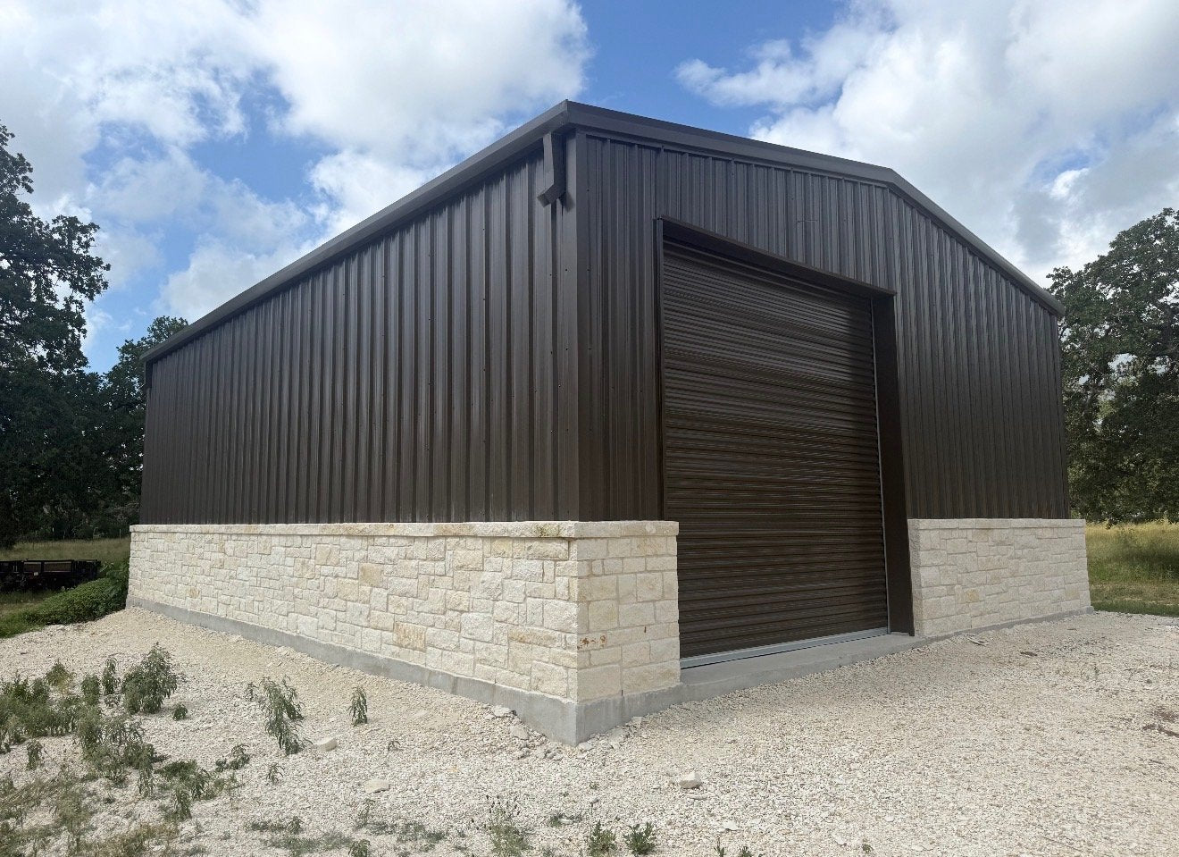 Custom metal workshop building with bronze siding, limestone wainscot, and a wide roll-up bay door under Texas oak trees