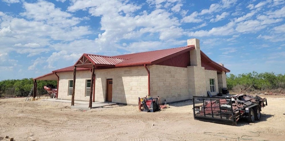 Limestone custom home with a red standing seam metal roof and covered porch, under construction on a cleared West Texas lot
