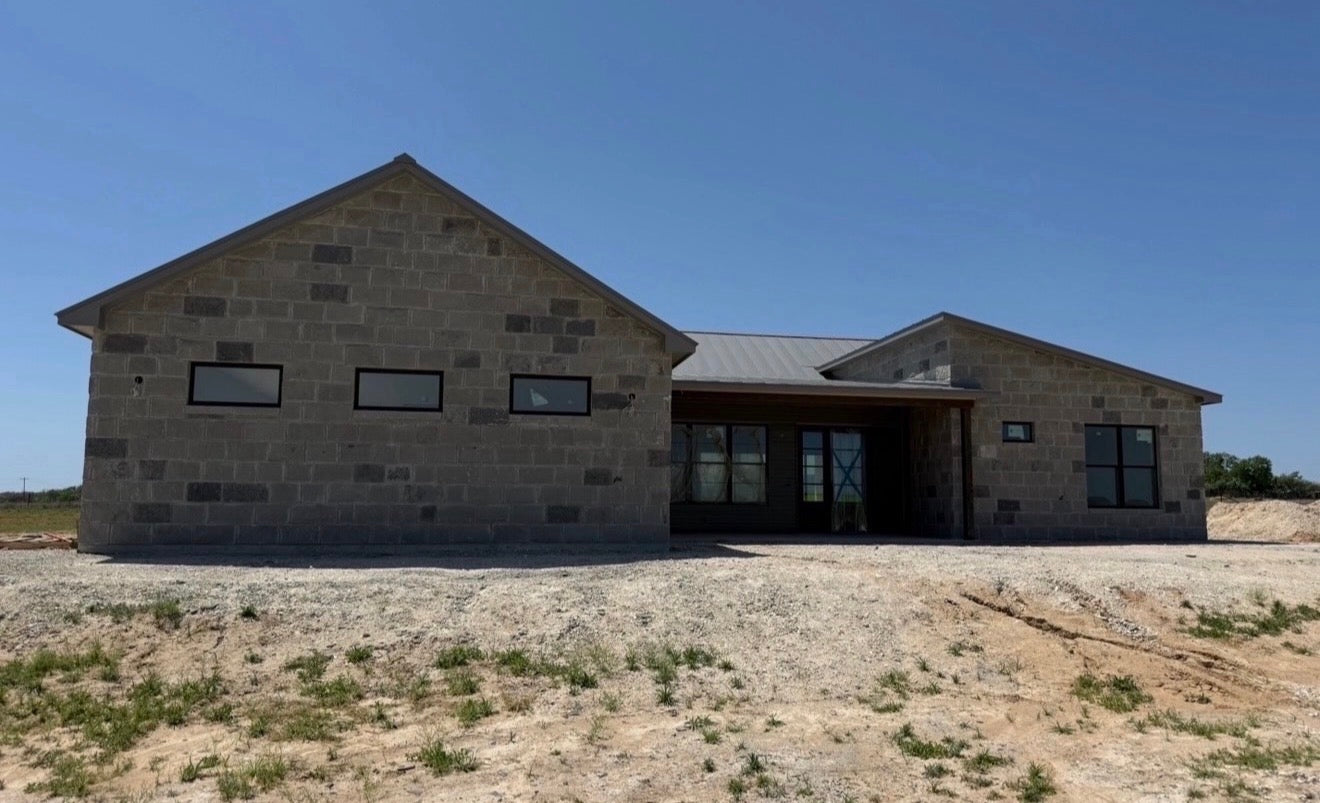 Front elevation of a single-story limestone custom home with a dark metal roof and recessed entry on open Texas land