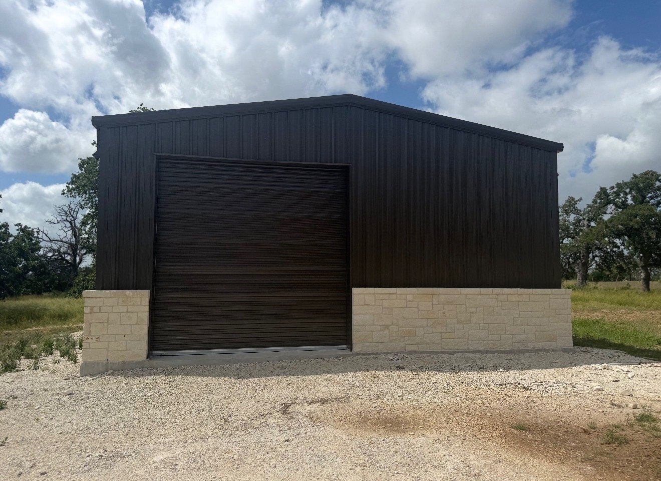 Front elevation of a black metal shop building with an oversized roll-up door and limestone base on a Texas property
