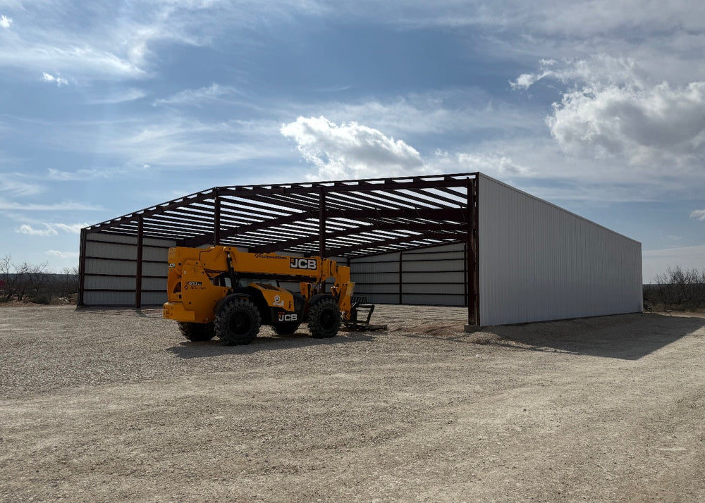 Red-iron steel building frame with partial wall sheeting installed and a telehandler parked inside the open bay
