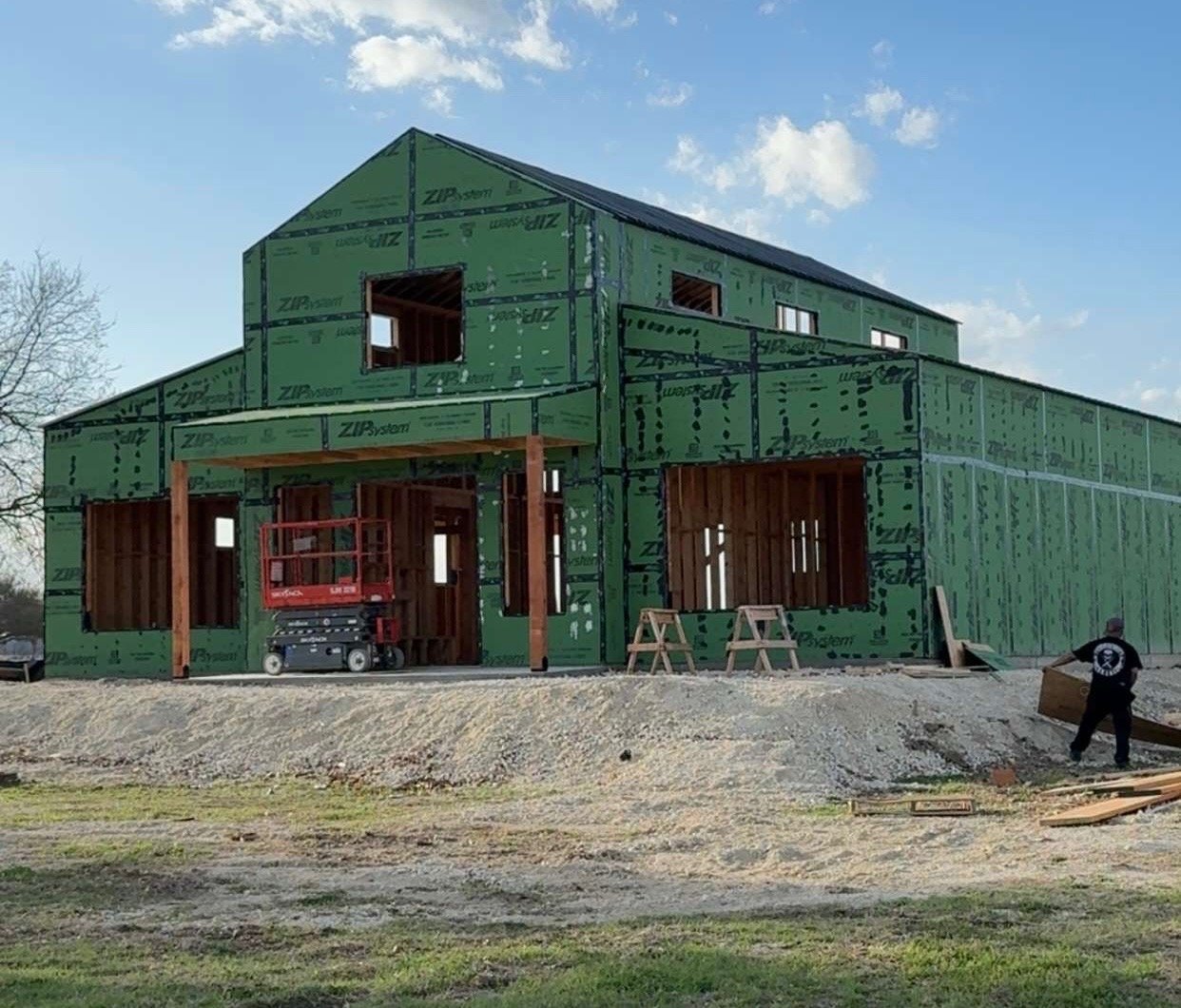 Two-story barndominium under construction with ZIP System sheathing, timber porch posts, and a scissor lift on site