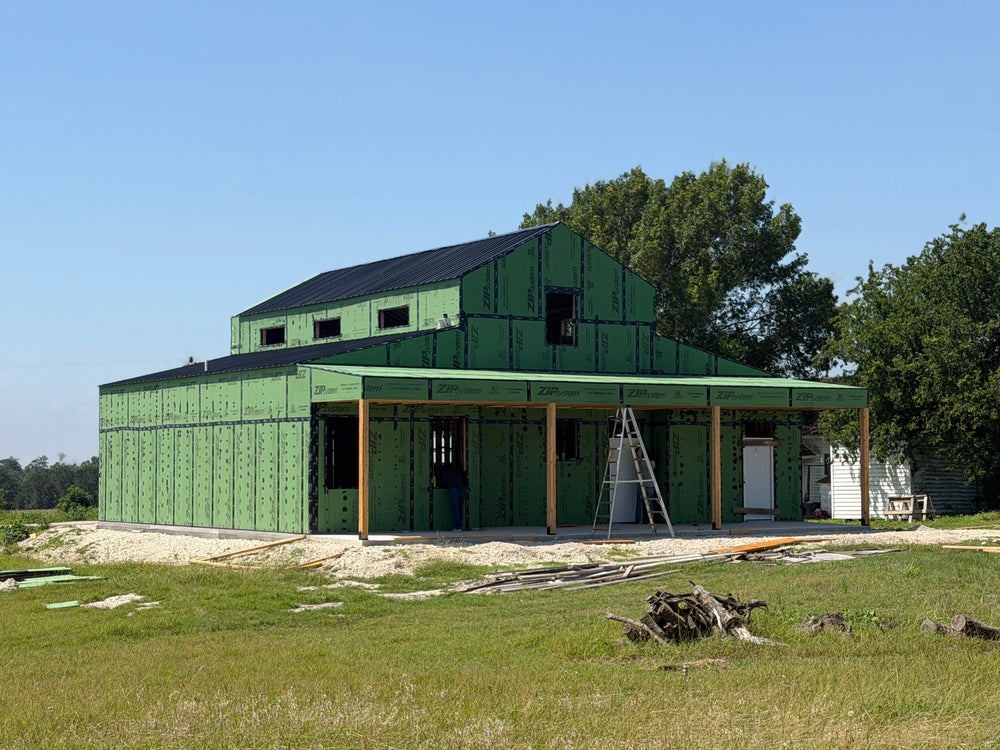 Two-story barndominium under construction with green ZIP System sheathing, a black metal roof, and timber posts framing the wraparound porch
