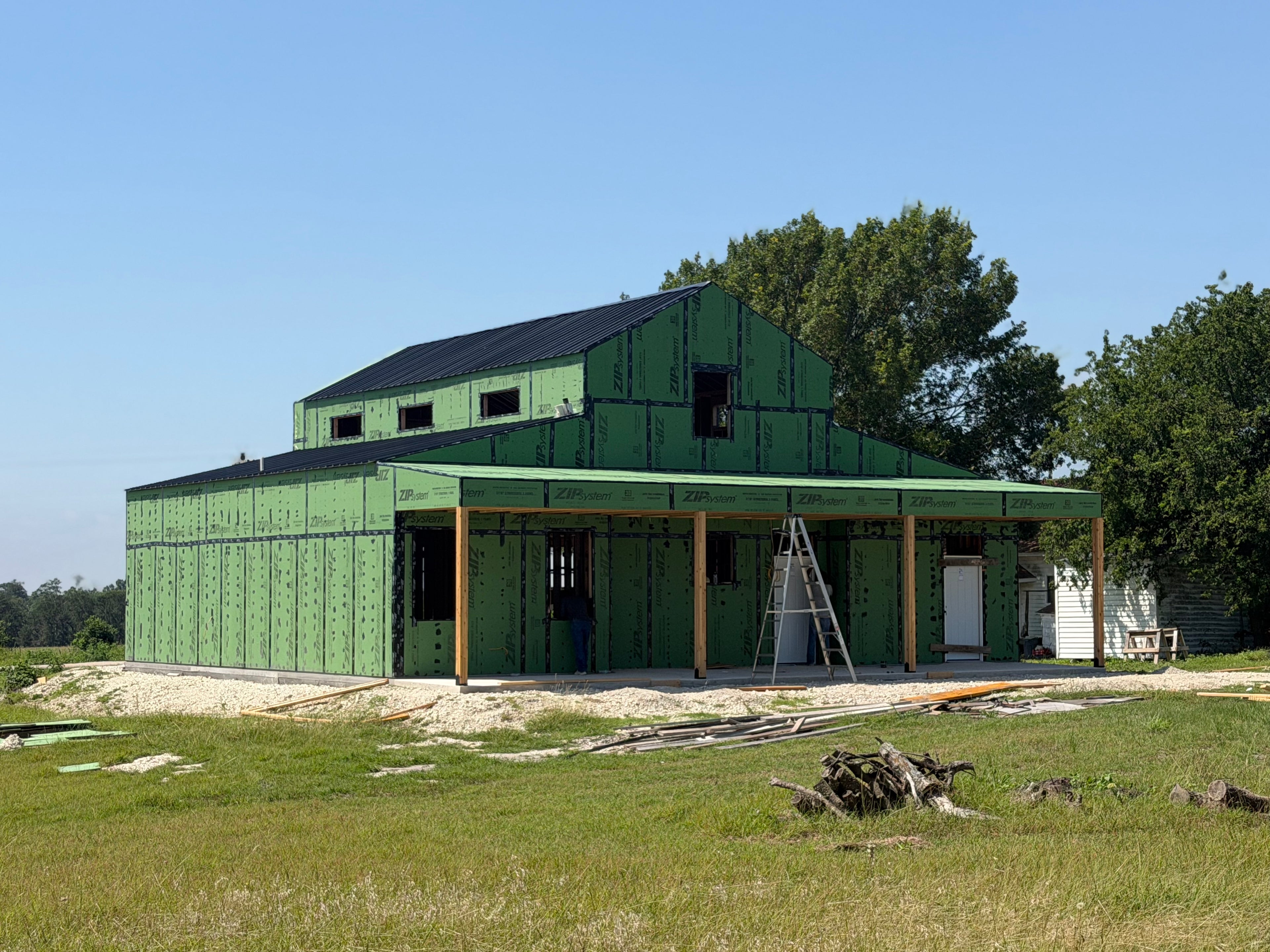 Two-story barndominium under construction with green ZIP System sheathing, a black metal roof, and timber posts framing the wraparound porch