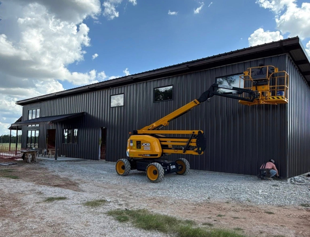 Two-story dark metal barndominium under construction with an articulating boom lift positioned for upper-level siding work