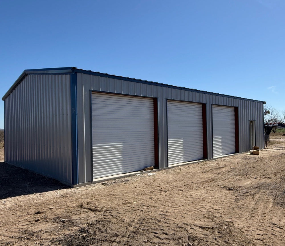 White metal garage building with three roll-up doors on a graded dirt pad
